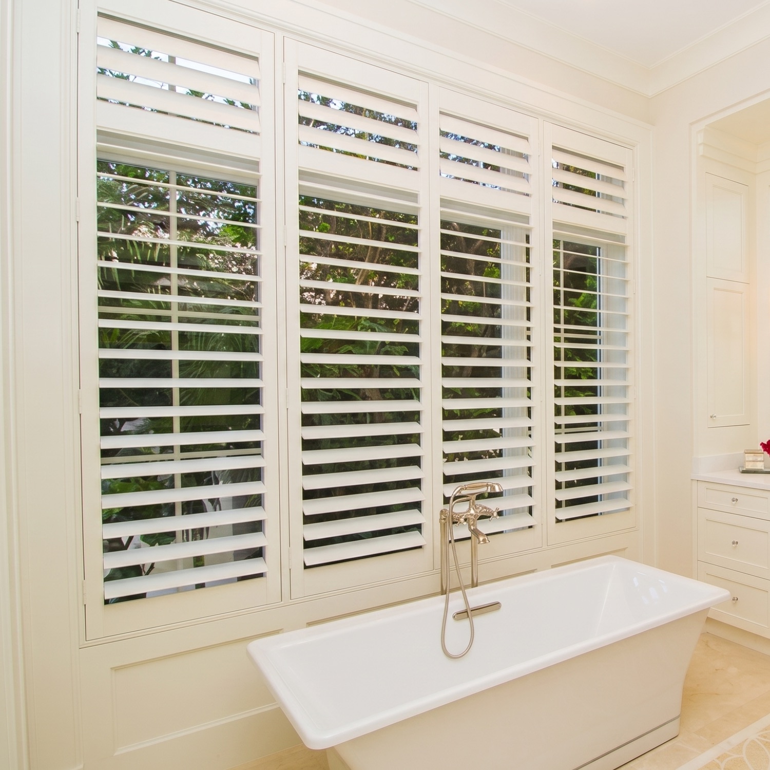 White hardwood plantation shutters in a Naples bathroom with freestanding tub and tropical garden views